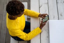 D'en haut de la messagerie féminine afro-américaine concentrée avec smartphone tout en se relaxant à la table en bois — Photo de stock
