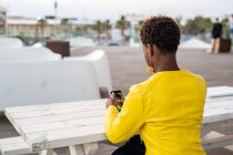 Vue arrière de la messagerie féminine afro-américaine concentrée avec smartphone tout en se relaxant à la table en bois — Photo de stock