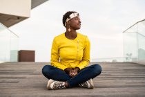 Стильна афроамериканець жінка в сучасній куртці розслабляючий сидячи на дерев'яній підлозі і оглядаючись — стокове фото