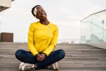 Stylish African American female in modern jacket relaxing sitting on wooden floor and looking in camera making silly faces — Stock Photo