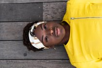From above stylish African American female in modern jacket relaxing on wooden floor and looking in camera — Stock Photo