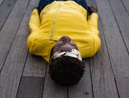 Сверху стильная афроамериканка в современной куртке отдыхает на деревянном полу и смотрит в сторону — стоковое фото