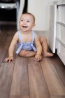 Bébé pieds nus excité regardant loin tout en étant assis sur le parquet près des comptoirs dans une cuisine confortable à la maison — Photo de stock