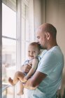 Vista lateral do homem careca abraçando e beijando o bebê feliz enquanto está perto da janela no quarto acolhedor em casa — Fotografia de Stock