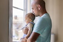 Вид сбоку лысого мужчины, который обнимает и целует счастливого ребенка, стоя у окна в уютной комнате дома — стоковое фото