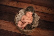Nude infant in knitted hat sleeping in bucket on wooden floor at home — Stock Photo