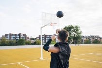 Young man throwing ball while playing on basketball court outdoors in rear view. — Stock Photo