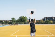 Young man throwing black ball on yellow basketball court outdoors in rear view. — Stock Photo