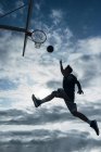 Young man jumping for scoring on basketball court outdoors against cloudy sky. — Stock Photo