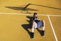 Jugador de baloncesto masculino usando smartphone mientras está acostado en la cancha amarilla después de la sesión de entrenamiento . - foto de stock