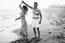 Adult man smiling and spinning woman in dance while having fun on sandy beach near sea — Stock Photo