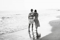 Rückansicht des barfüßigen Mannes und der barfüßigen Frau, die sich umarmen, während sie am Sandstrand in Richtung des winkenden Meeres am Urlaubsort gehen — Stockfoto