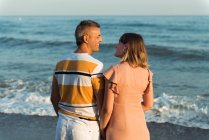 Hombre y mujer adultos de pie en la playa cerca del mar ondeando y mirándose el uno al otro - foto de stock