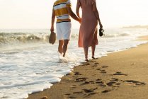 Back view of barefoot man and woman holding hands and carrying shoes while walking on sandy beach — Stock Photo