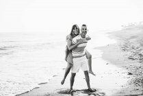 Full length cheerful adult male giving piggyback ride to smiling female while standing on wet sand — Stock Photo