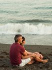 Взрослый мужчина и женщина в полной длине улыбаются и обнимаются, сидя на песке возле моря и расслабляясь во время свидания — стоковое фото