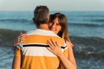 Adult man hugging woman while standing on beach near waving sea and resting together — Stock Photo