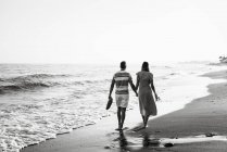 Vista trasera del hombre y la mujer descalzos tomados de la mano y llevando zapatos mientras caminan en la playa de arena - foto de stock