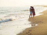 Back view of barefoot man and woman kissing while walking on sandy beach towards waving sea on resort — Stock Photo
