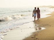Rückansicht des barfüßigen Mannes und der barfüßigen Frau, die sich beim Spazierengehen am Sandstrand an den Händen halten und Schuhe tragen — Stockfoto