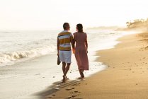 Rückansicht des barfüßigen Mannes und der barfüßigen Frau, die sich beim Spazierengehen am Sandstrand an den Händen halten und Schuhe tragen — Stockfoto
