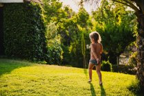 Повне тіло без сорочки маленька дівчинка в трусиках грає під краплями чистої води, розважаючись на газоні у дворі в сонячний літній день — стокове фото