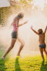 Ganzkörperanonymer Teenager und kleines Mädchen rennen und spielen in Tropfen, die sauberes Wasser versprühen, während sie gemeinsam Spaß im Garten haben — Stockfoto