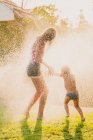 Полностью анонимный подросток и маленькая девочка бегают и играют в капли брызг чистой воды, веселясь в саду вместе — стоковое фото