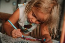 Konzentriertes kleines Mädchen lehnt am Tisch und malt Bilder am Buch mit Filzstift auf verschwommenem Hintergrund des Zimmers zu Hause aus — Stockfoto