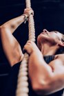 From below muscular woman climbing rope and looking up on black background - foto de stock