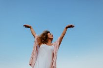 Низкий угол элегантной женщины с вьющимися волосами закрывая глаза и поднимая руки стоя против безоблачного голубого неба в природе — стоковое фото