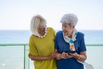 Старые друзья в элегантных нарядах вместе просматривают смартфон, стоя и отдыхая на балконе напротив моря на курорте — стоковое фото