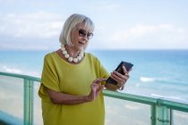 Elegante mujer mayor navegando por las redes sociales en el teléfono inteligente y mirando hacia otro lado mientras está de pie en el balcón - foto de stock