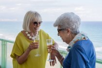 Mulheres felizes em roupas elegantes dizendo brinde e desfrutando de vinho enquanto estão de pé na varanda do hotel perto do mar no resort — Fotografia de Stock