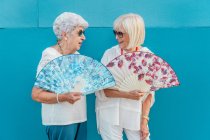 Positivas fêmeas de cabelos grisalhos envelhecidas na moda acenando com grandes ventiladores de mão coloridos, olhando um para o outro no fundo azul — Fotografia de Stock