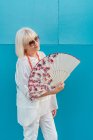 Elegant elderly woman with fan smiling and looking at camera while standing against blue wall on hot day on resort — Stock Photo