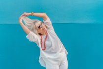 Портрет красивой беловолосой пожилой женщины в солнечных очках в белой рубашке на фоне синей стены — стоковое фото