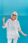 Portrait of beautiful stylish white-haired senior woman with sunglasses in white shirt with coral beads on blue wall background — Stock Photo