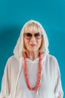 Porträt der schönen eleganten weißhaarigen Seniorin mit Sonnenbrille im weißen Hemd mit Kapuze auf dem Kopf und Korallenperlen — Stockfoto