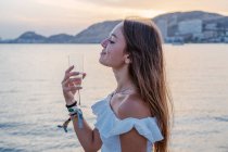 Вид сбоку счастливой молодой женщины с закрытыми глазами, пьющей вино, стоя у моря вечером на курорте — стоковое фото