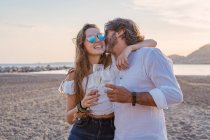 Зрелый мужчина обнимает и целует молодую женщину в щеку, предлагая тосты и празднуя воссоединение семьи на песчаном пляже во время заката на курорте — стоковое фото