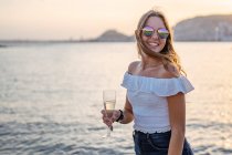 Jovem mulher feliz com um copo de vinho enquanto está perto do mar à noite no resort — Fotografia de Stock