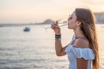 Vista laterale di felice giovane femmina con gli occhi chiusi bere vino mentre in piedi vicino al mare in serata in resort — Foto stock