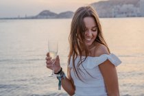 Feliz joven hembra con una copa de vino mientras está de pie cerca del mar por la noche en el complejo - foto de stock