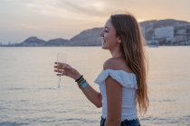 Felice giovane femmina con un bicchiere di vino mentre in piedi vicino al mare in serata in resort — Foto stock