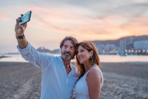 Бородатий зрілий чоловік бере селфі з дружиною, проводячи на піщаному пляжі під час заходу сонця разом — стокове фото
