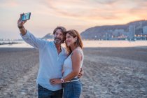 Barbu homme mûr prendre selfie avec femme tout en passant sur la plage de sable au coucher du soleil ensemble — Photo de stock