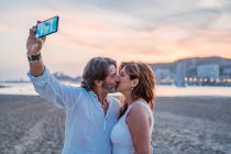 Barbu homme mûr prendre selfie avec femme tout en passant sur la plage de sable au coucher du soleil ensemble — Photo de stock