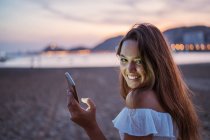 Vista trasera de la joven y feliz mujer sonriendo y navegando por las redes sociales en el teléfono inteligente mientras pasa tiempo en la playa de arena por la noche - foto de stock