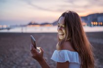 Back view of happy young female smiling and browsing social media on smartphone while spending time on sandy beach in evening — Stock Photo